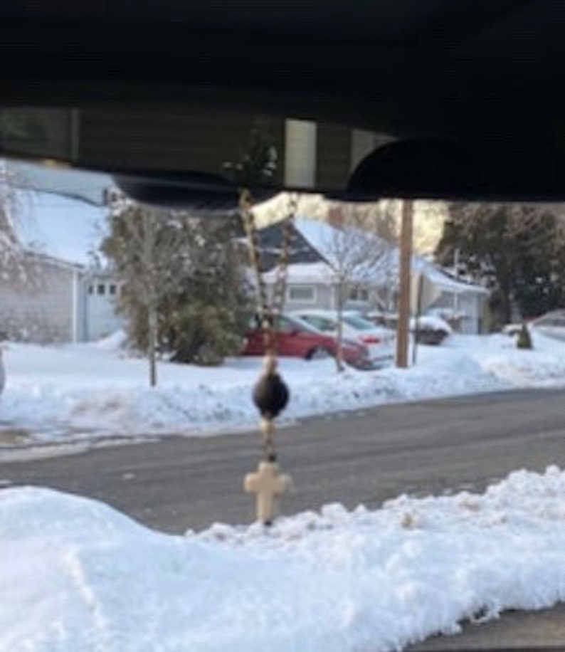 May include: A hanging rosary ornament featuring a gold-toned metal chain, a single large black spherical bead, and a small off-white cross pendant. The religious item is suspended from a vehicle rearview mirror, displaying a simple design.