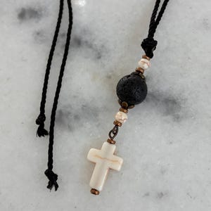 May include: A necklace featuring a black cord, a large porous black lava bead, smaller white marbled beads with brown veining, and a white cross pendant. The components are connected by copper-toned metal spacers and findings.