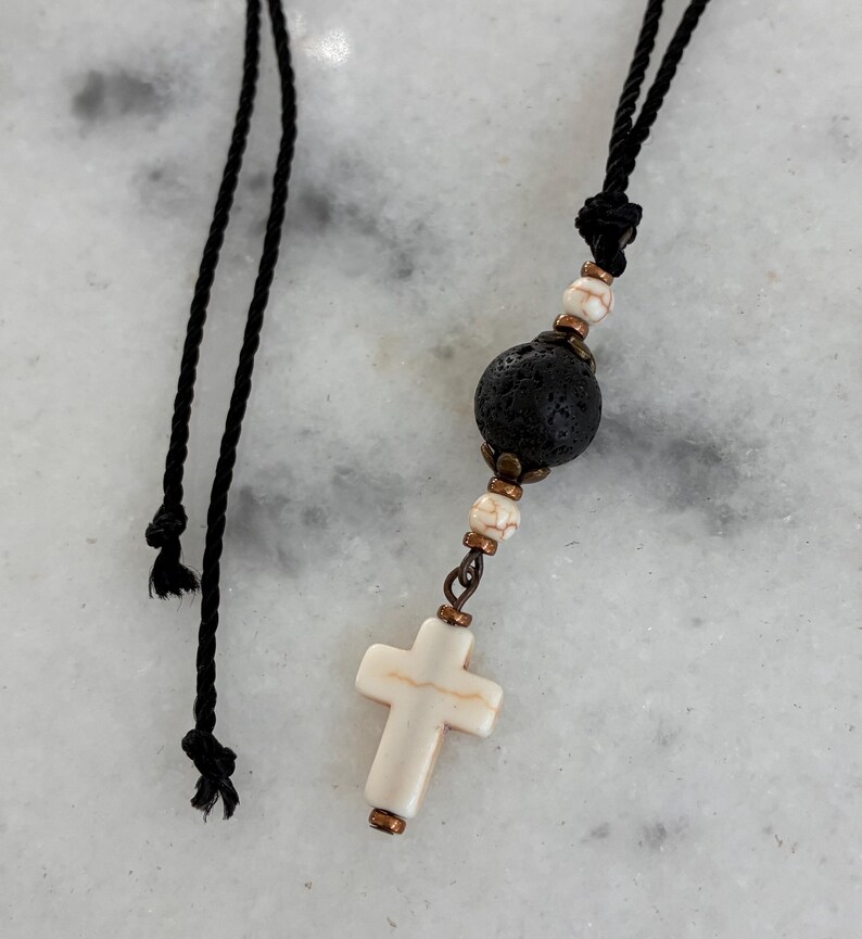 May include: A necklace featuring a black cord, a large porous black lava bead, smaller white marbled beads with brown veining, and a white cross pendant. The components are connected by copper-toned metal spacers and findings.