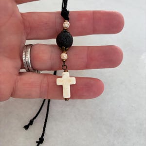May include: A necklace featuring a black cord, a central porous black lava bead, two smaller white marbled beads, and a white cross pendant. The beads are accented with small bronze-toned metal caps and connectors, creating a layered design.