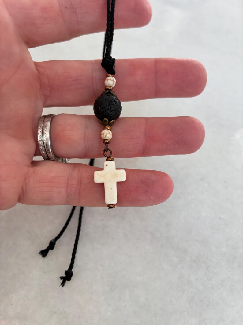 May include: A necklace featuring a black cord, a central porous black lava bead, two smaller white marbled beads, and a white cross pendant. The beads are accented with small bronze-toned metal caps and connectors, creating a layered design.