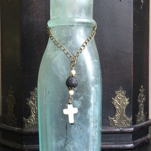 May include: A light blue glass bottle adorned with a bronze-toned chain necklace featuring a black porous lava bead, two small cream-colored spherical beads, and a white cross-shaped pendant hanging from the center of the chain.