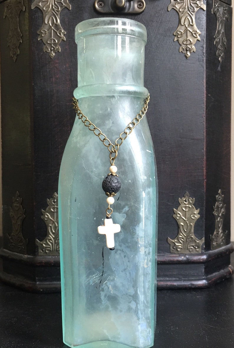 May include: A light blue glass bottle adorned with a bronze-toned chain necklace featuring a black porous lava bead, two small cream-colored spherical beads, and a white cross-shaped pendant hanging from the center of the chain.
