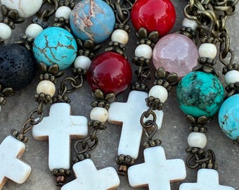 May include: Close-up of multiple necklaces with white crosses and colorful beads. The beads are various colors, including red, turquoise, and white. The chains are a bronze color, and the crosses are white.