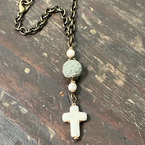 May include: A necklace featuring a bronze-toned metal chain with a lobster clasp, a central porous grey lava stone bead, two small white beads, and a white cross-shaped pendant suspended at the bottom of the chain assembly.
