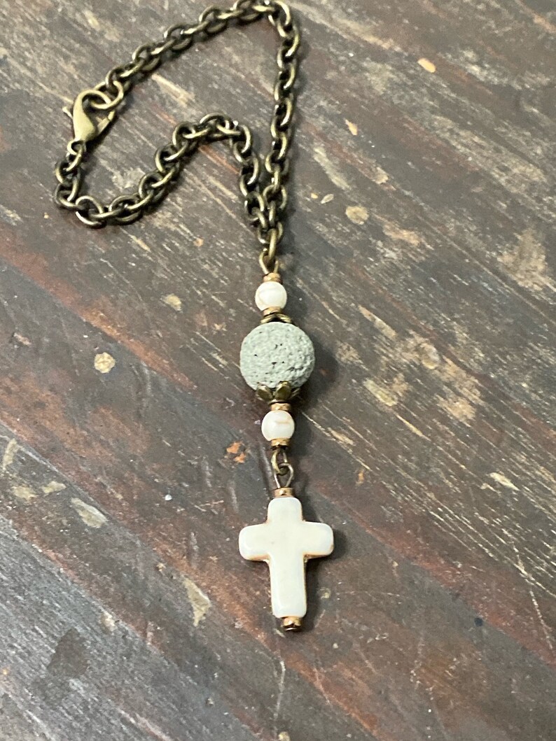 May include: A necklace featuring a bronze-toned metal chain with a lobster clasp, a central porous grey lava stone bead, two small white beads, and a white cross-shaped pendant suspended at the bottom of the chain assembly.