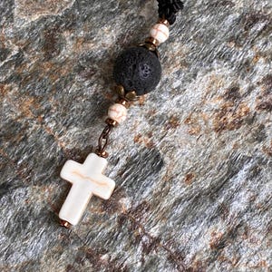 May include: A necklace featuring a black cord, a large porous black lava bead, two smaller white beads with brown veining, and a white cross pendant with brown veining. The beads are accented with small metallic bronze-toned spacers and caps.