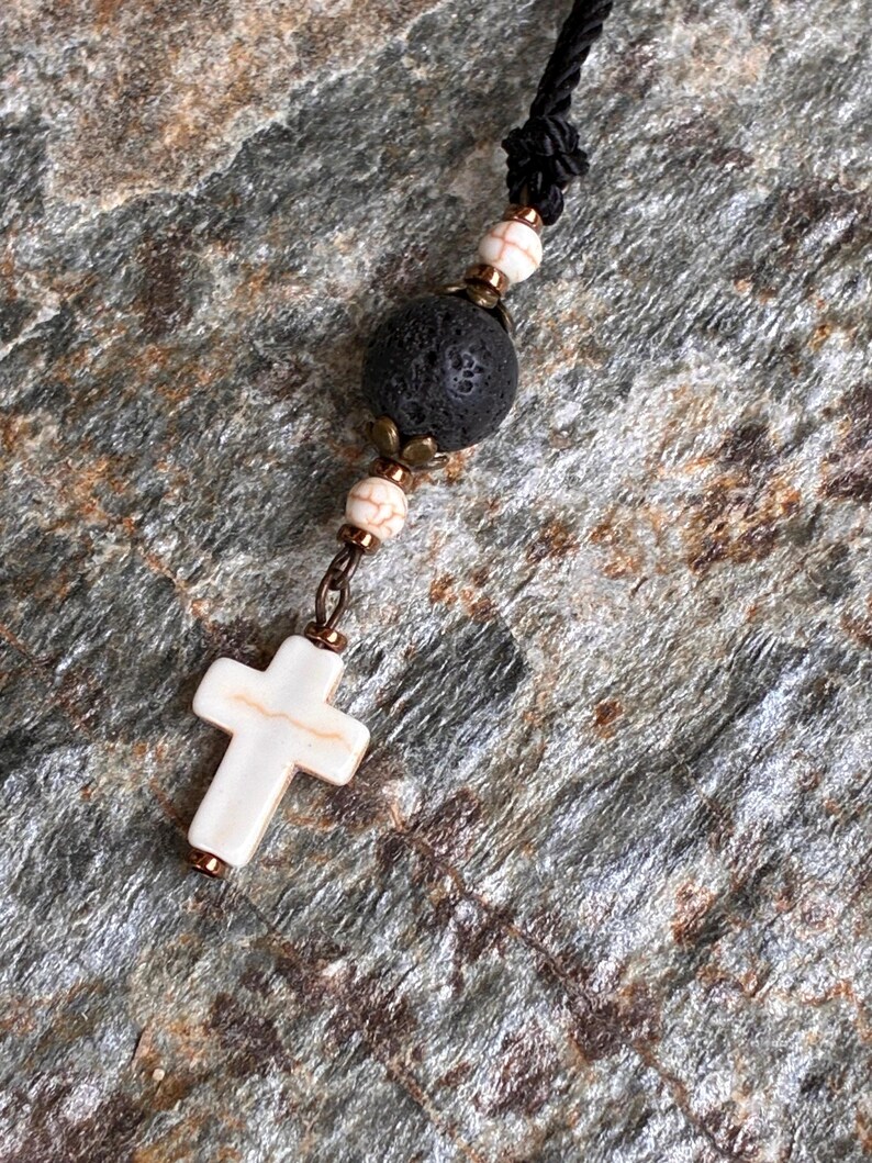 May include: A necklace featuring a black cord, a large porous black lava bead, two smaller white beads with brown veining, and a white cross pendant with brown veining. The beads are accented with small metallic bronze-toned spacers and caps.