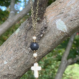 May include: A necklace featuring a bronze-toned metal chain, a central black porous lava bead, two small white round beads, and a white cross-shaped pendant. The beads are separated by small bronze-toned metal spacers, creating a vertical arrangement.