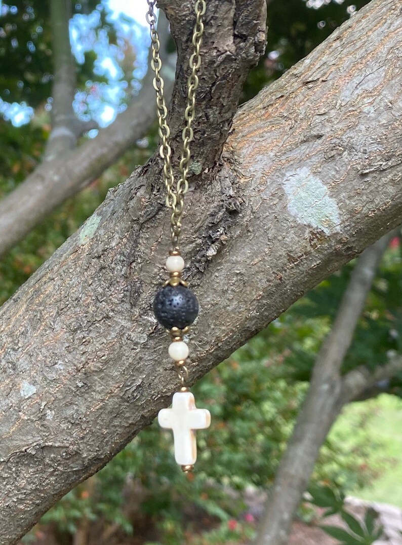May include: A necklace featuring a bronze-toned metal chain, a central black porous lava bead, two small white round beads, and a white cross-shaped pendant. The beads are separated by small bronze-toned metal spacers, creating a vertical arrangement.