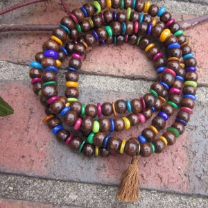 May include: A multi-strand mala necklace with dark brown wooden beads and colorful accent beads in blue, green, yellow, pink, and orange. A brown tassel hangs from the bottom. The necklace is displayed on a brick surface.