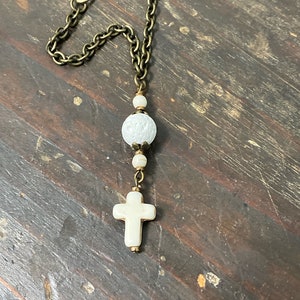 May include: A decorative hanging ornament featuring a bronze-toned metal chain with a lobster clasp. The chain connects to a series of white beads, including one large textured spherical bead and a smooth cross-shaped pendant at the bottom.
