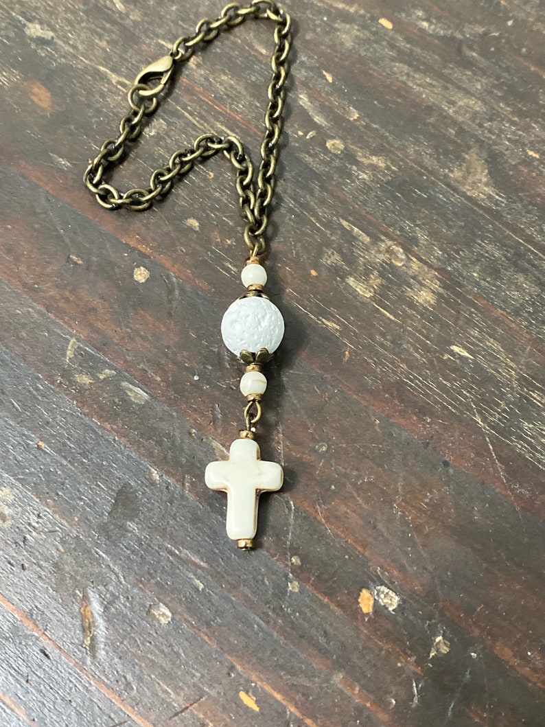 May include: A decorative hanging ornament featuring a bronze-toned metal chain with a lobster clasp. The chain connects to a series of white beads, including one large textured spherical bead and a smooth cross-shaped pendant at the bottom.