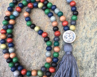 May include: A multi-colored wooden bead necklace with a silver Om charm and a gray tassel. The beads are in shades of green, blue, red, brown, and black.