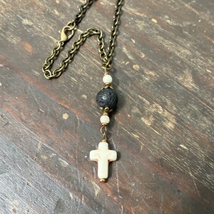 May include: A necklace featuring an antique bronze-toned metal chain with a lobster clasp. The pendant consists of a central black porous lava bead flanked by two small white beads, terminating in a white cross-shaped charm with bronze accents.