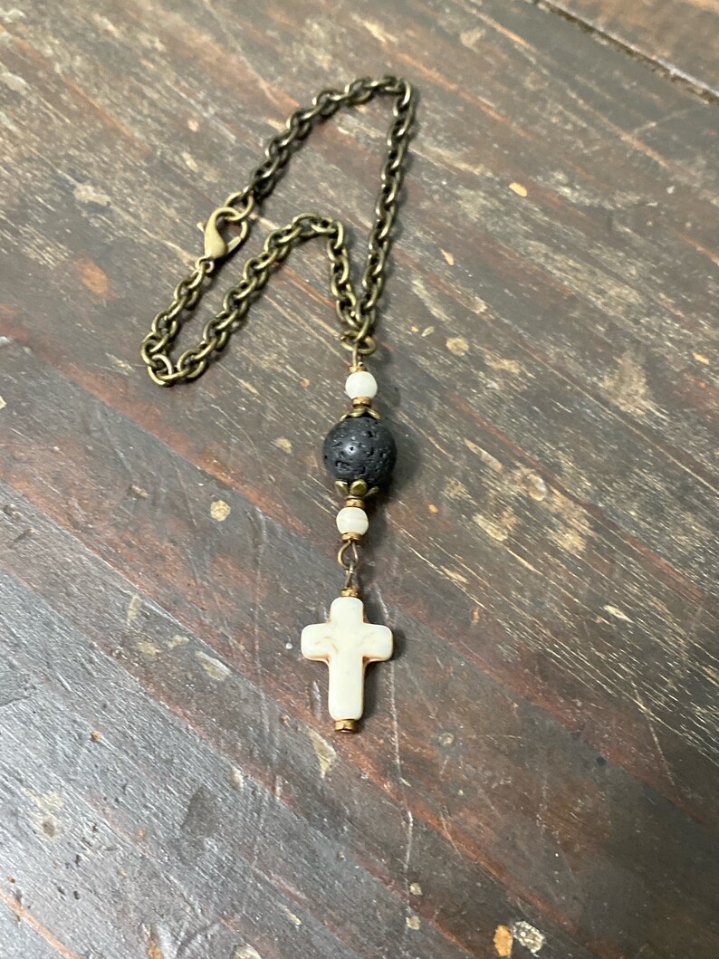 May include: A necklace featuring an antique bronze-toned metal chain with a lobster clasp. The pendant consists of a central black porous lava bead flanked by two small white beads, terminating in a white cross-shaped charm with bronze accents.