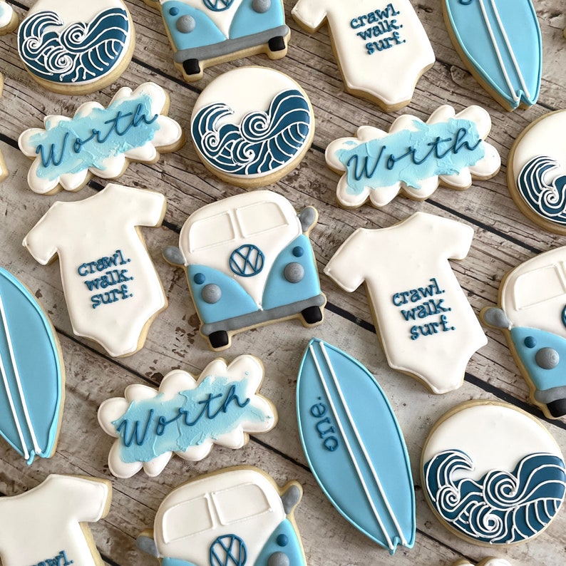 Pode incluir: Um conjunto de 12 bolachas decoradas em azul e branco com v&aacute;rios desenhos, incluindo uma prancha de surf, ondas, uma van Volkswagen e um macac&atilde;o com o texto "crawl, walk, surf". As bolachas s&atilde;o decoradas com glac&ecirc; azul e branco e t&ecirc;m a palavra "Worth" escrita em v&aacute;rias delas.