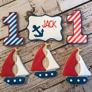 May include: A set of nautical themed sugar cookies decorated with red, white, and blue icing. The set includes two cookies shaped like the number one, one cookie with a sign that says "JACK" with an anchor, and three cookies shaped like sailboats.