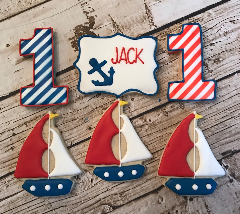 May include: A set of nautical themed sugar cookies decorated with red, white, and blue icing. The set includes two cookies shaped like the number one, one cookie with a sign that says "JACK" with an anchor, and three cookies shaped like sailboats.
