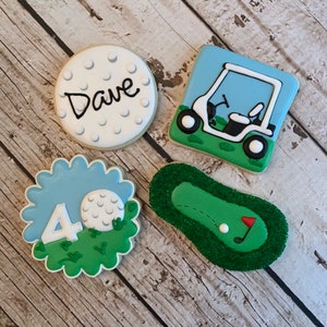 May include: Four decorated sugar cookies with a golf theme. One cookie is a golf ball with the name "Dave" written on it. Another cookie is a golf ball on a green with the number "40" in front of it. A third cookie is a golf cart on a green. The last cookie is a green with a golf ball and a flag.