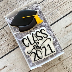 May include: Two decorated sugar cookies in a white box with white shredded paper. One cookie is black and shaped like a graduation cap with a yellow tassel. The other cookie is white with black icing that says "Class of 2021".
