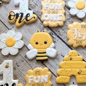 May include: A set of ten decorated sugar cookies featuring a bee theme. The cookies are decorated with yellow, white, and black icing. The cookies feature a beehive, bees, flowers, and the words "It's Fun To Bee One".