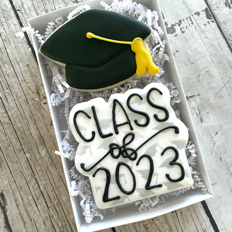 May include: A white cookie decorated with black icing that says "Class of 2023" and a small black and yellow graduation cap cookie.