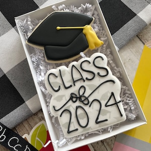 May include: Two decorated sugar cookies in a white box. One cookie is a black graduation cap with a yellow tassel. The other cookie is white with black lettering that says "Class of 2024" and a pattern of small black graduation caps.