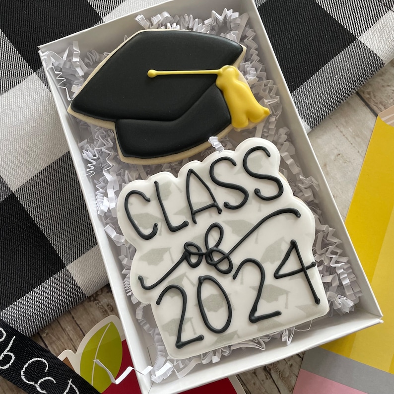 May include: Two decorated sugar cookies in a white box. One cookie is a black graduation cap with a yellow tassel. The other cookie is white with black lettering that says "Class of 2024" and a pattern of small black graduation caps.