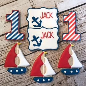 May include: Set of nautical themed sugar cookies decorated with red, white, and blue icing. The cookies include two sailboats, two number one cookies, and two cookies with the name "Jack" and an anchor.