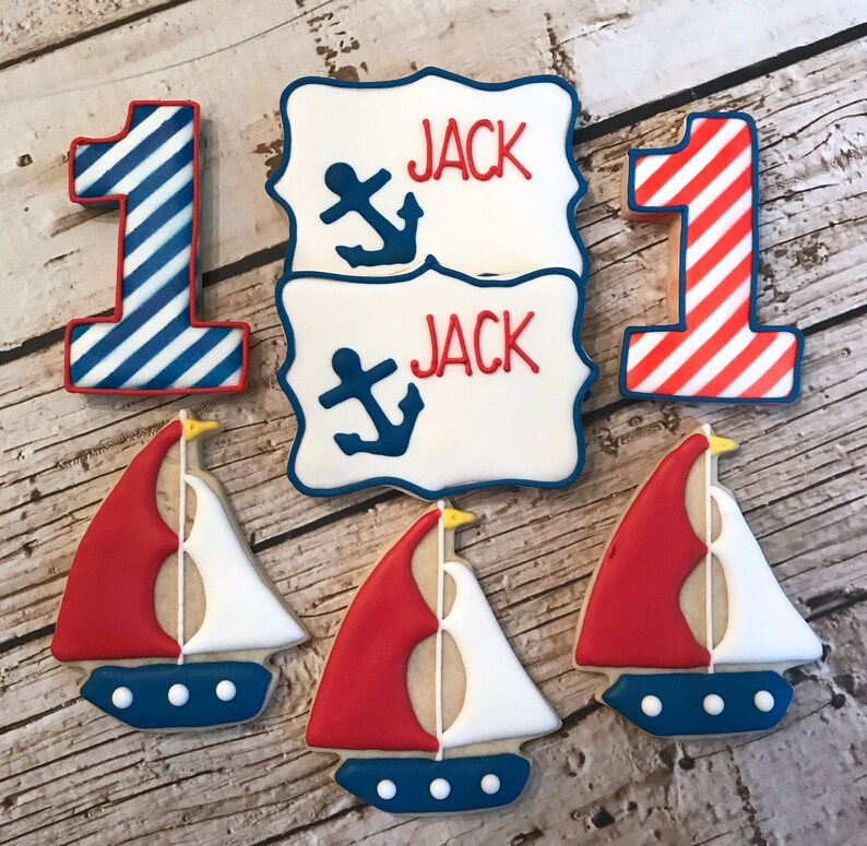 May include: Set of nautical themed sugar cookies decorated with red, white, and blue icing. The cookies include two sailboats, two number one cookies, and two cookies with the name "Jack" and an anchor.