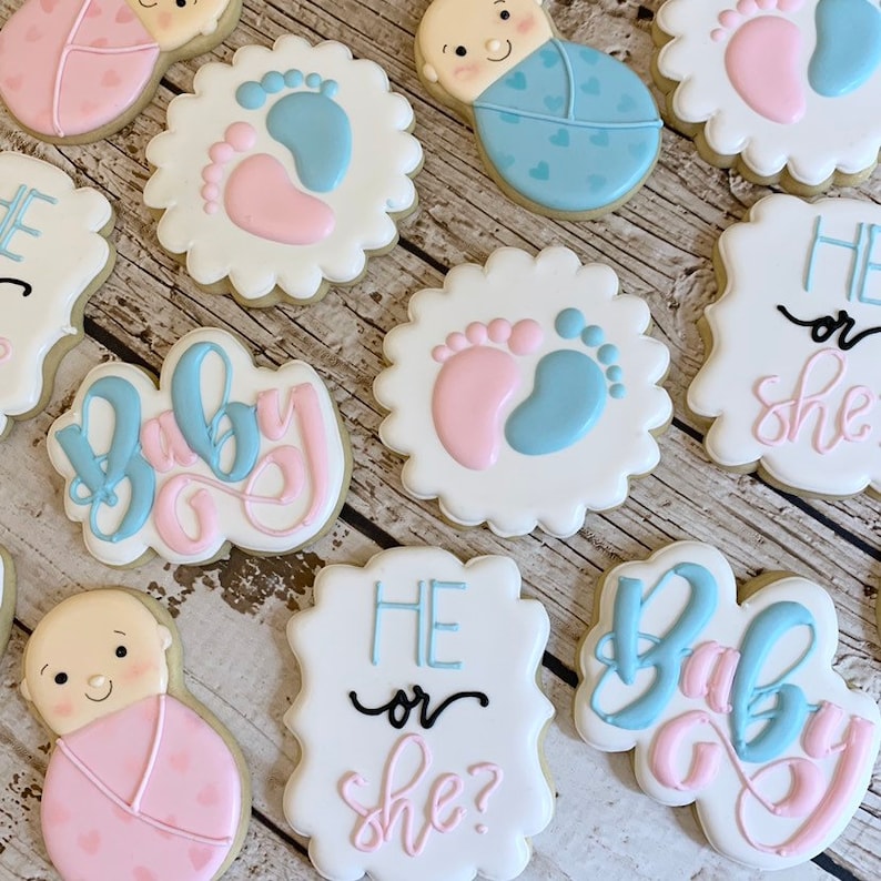 May include: A set of nine decorated sugar cookies with blue and pink frosting. The cookies feature baby footprints, a baby in a swaddle, and the text "He or She?" and "Baby Boy".