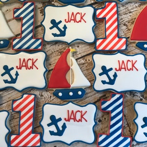 May include: A set of nine decorated sugar cookies with a nautical theme. The cookies are decorated with red, white, and blue icing. Some cookies are shaped like the number one, others are shaped like sailboats, and others are rectangular with the name "Jack" and an anchor design.