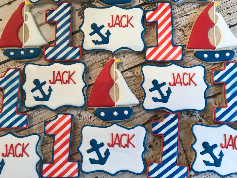 May include: A set of nine decorated sugar cookies with a nautical theme. The cookies are decorated with red, white, and blue icing. Some cookies are shaped like the number one, others are shaped like sailboats, and others are rectangular with the name "Jack" and an anchor design.