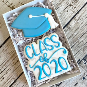 May include: A white gift box with blue and white decorated sugar cookies. The cookies are shaped like a graduation cap and a banner that says "Class of 2020".