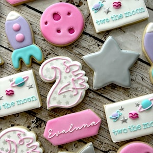 May include: A set of decorated sugar cookies with a space theme. The cookies feature a rocket, stars, planets, and the words "two the moon" and "Evalunna".