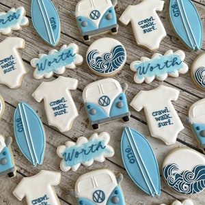 Pode incluir: Um conjunto de 12 bolachas de a&ccedil;&uacute;car decoradas com um esquema de cores azul e branco. As bolachas t&ecirc;m o formato de pranchas de surf, bodies de beb&eacute; e furgonetas Volkswagen. As bolachas s&atilde;o decoradas com as palavras "Worth", "One" e "Crawl, Walk, Surf".