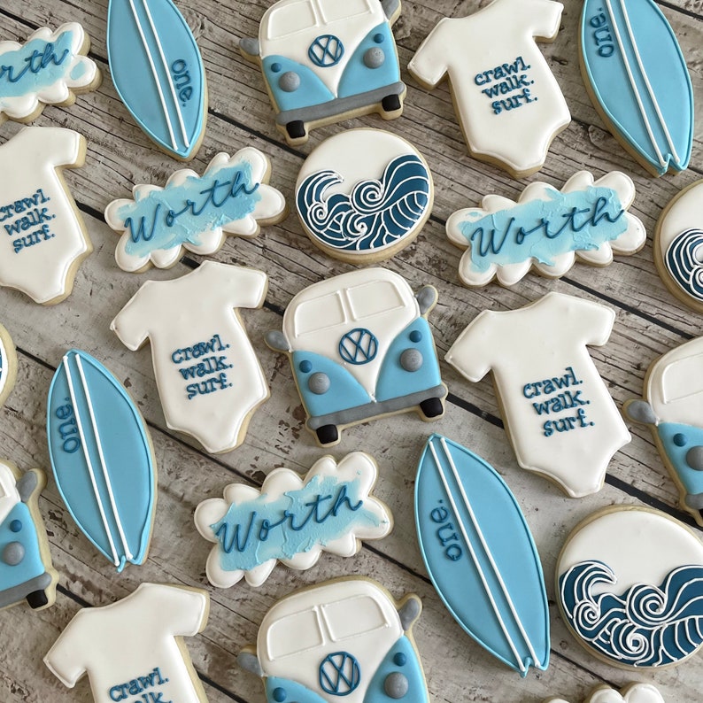 Pode incluir: Um conjunto de 12 bolachas de a&ccedil;&uacute;car decoradas com um esquema de cores azul e branco. As bolachas t&ecirc;m o formato de pranchas de surf, bodies de beb&eacute; e furgonetas Volkswagen. As bolachas s&atilde;o decoradas com as palavras "Worth", "One" e "Crawl, Walk, Surf".
