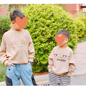 May include: Two children wearing beige sweatshirts. The child on the left is wearing blue jeans with a black heart patch and the text "NEVERULE" on their sweatshirt. The child on the right is wearing black and white striped pants and a sweatshirt with the text "MICKEY" and a graphic of Mickey Mouse and his friends.
