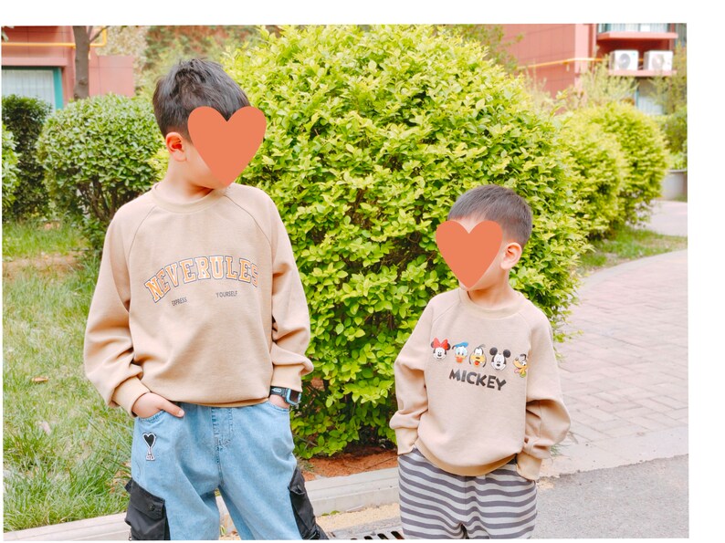 May include: Two children wearing beige sweatshirts. The child on the left is wearing blue jeans with a black heart patch and the text "NEVERULE" on their sweatshirt. The child on the right is wearing black and white striped pants and a sweatshirt with the text "MICKEY" and a graphic of Mickey Mouse and his friends.