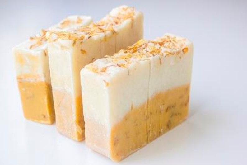 Puede incluir: Tres barras rectangulares de jab&oacute;n artesanal. La mitad superior de cada barra es blanca, pasando a un tono amarillo-naranja en la parte inferior. La parte superior de cada barra est&aacute; salpicada de peque&ntilde;os copos de color marr&oacute;n claro. Los jabones est&aacute;n dispuestos sobre una superficie blanca.