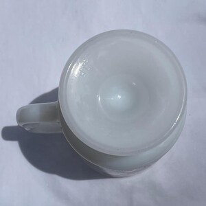May include: A white, opaque glass mug with a rounded body and a small handle. The mug's interior has a conical shape. The mug is sitting on a white surface. The mug is empty.