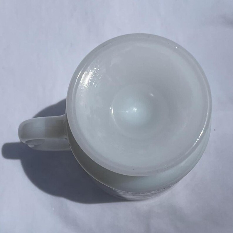 May include: A white, opaque glass mug with a rounded body and a small handle. The mug's interior has a conical shape. The mug is sitting on a white surface. The mug is empty.