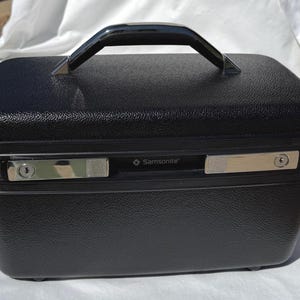 May include: A black Samsonite vanity case with a textured exterior. The case has a black handle and two silver latches. The Samsonite logo is visible on the front of the case. The case is rectangular and appears to be made of a durable material.