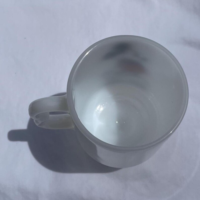 May include: A white ceramic mug with a rounded body and a curved handle. The mug is empty and appears to be clean. The mug casts a shadow on the white surface it sits on.