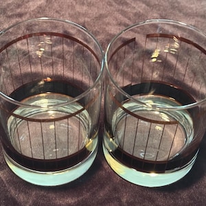 May include: Two clear, vintage-style drinking glasses with a brown band and vertical lines design. The glasses have a low, rounded base and are filled with a clear liquid. The glasses are set against a dark purple background.