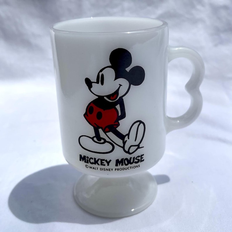 May include: White glass mug featuring Mickey Mouse in a classic pose, with black ears, a red shorts, and white gloves and shoes. The mug has a heart-shaped handle and the text "Mickey Mouse" and "Walt Disney Productions".