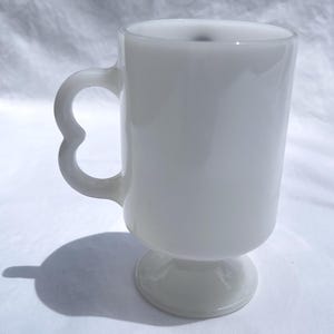 May include: A white, opaque glass mug with a heart-shaped handle. The mug has a pedestal base and a cylindrical body. The handle is a unique design element. The mug is likely vintage and suitable for hot beverages.
