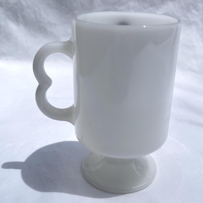 May include: A white, opaque glass mug with a heart-shaped handle. The mug has a pedestal base and a cylindrical body. The handle is a unique design element. The mug is likely vintage and suitable for hot beverages.