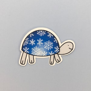 Può includere: Una tartaruga blu con un motivo a fiocco di neve bianco sul suo guscio. La tartaruga sorride e ha uno stile di cartone animato semplice.
